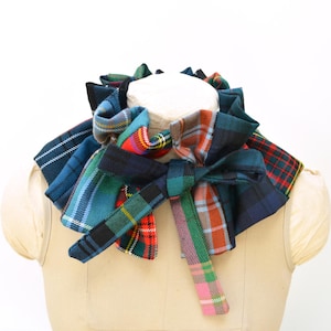 Puede incluir: Un accesorio para el cuello con volantes confeccionado con varios parches de tela de cuadros en tonos azul, verde, rojo y naranja. La pieza presenta un cierre central de lazo y un diseño voluminoso y superpuesto que rodea el cuello con pliegues.