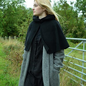 Model wearing black short cape made of fleece fabric. The cloak is worn over a grey wool coat and she is standing in front of a gate.