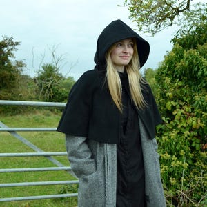 Woman wearing black fleece hooded short cape over a grey wool coat. She is wearing the cape with the hood up and is stood in front of a gate near a field.