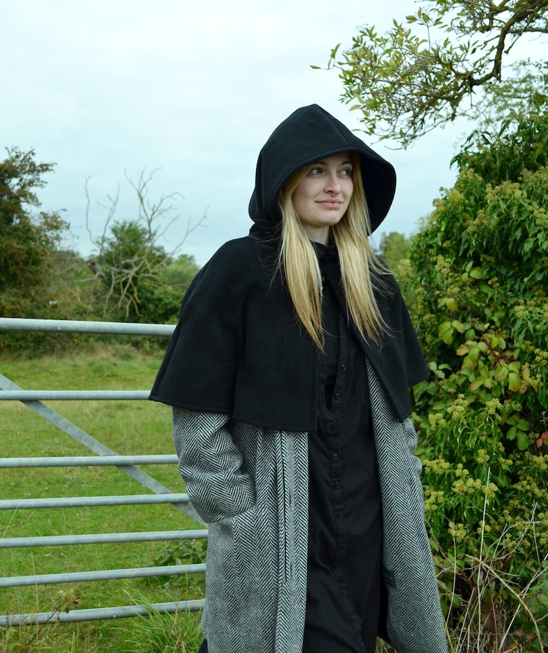 Woman wearing black fleece hooded short cape over a grey wool coat. She is wearing the cape with the hood up and is stood in front of a gate near a field.