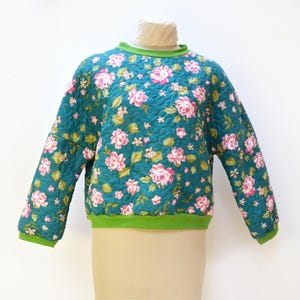 Puede incluir: Una sudadera acolchada de color verde azulado con un estampado floral de rosas rosas y hojas verdes. La sudadera tiene cuello y puños verdes. La prenda se exhibe en un maniquí.