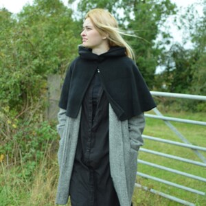 woman wearing a short black fleece cape, with the hood down. The mini cloak is worn over a grey coat and she is stood in a field.