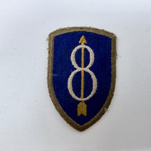 May include: A shield-shaped embroidered patch featuring a blue background with a tan border. Centered on the patch is a vertical gold arrow pointing upward, overlaid with a white figure-eight shape that encircles the shaft of the arrow.