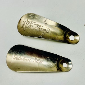 May include: Two vintage metal shoehorns with a tarnished finish. One shoehorn has the text "Walk-Over Shoe" with an illustration of a person. The other shoehorn has the text "Walk-Over Shoes". Both have a hole for hanging.