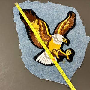 May include: Embroidered eagle patch with outstretched wings, featuring brown, yellow, and white details. The patch is on a piece of denim. A yellow measuring tape is placed across the patch, showing its size.