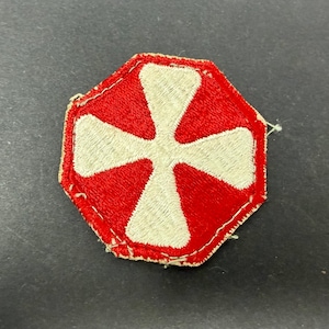 May include: A hexagonal embroidered patch featuring a red background with a white cross design. The edges of the patch are stitched with a light-colored thread, and the overall construction shows a dense, textured thread pattern throughout the surface.