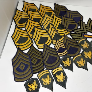 May include: A collection of vintage military patches, including chevrons and eagle insignia. The patches feature gold and yellow embroidery on dark blue, green, and black backgrounds. Some patches have a diamond shape in the center.