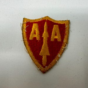 May include: Embroidered patch with a red shield shape, featuring the letters "AA" in gold, flanking a gold arrow. The patch has a gold border and is likely a military or organizational insignia.