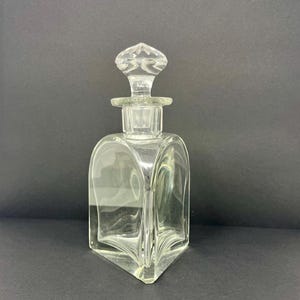 May include: A clear, vintage glass perfume bottle with a geometric design. The bottle has a square base that tapers to a point, with rounded sides and a decorative stopper. The stopper is diamond-shaped and sits atop a cylindrical neck.