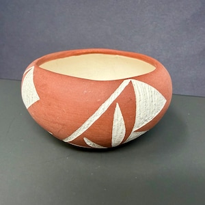 May include: A round, terracotta-colored ceramic bowl with a cream-colored interior. The exterior features a repeating pattern of white, angular shapes. The bowl is likely handmade and suitable for decorative or functional use.