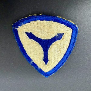 May include: A triangular embroidered patch featuring a tan background with a thick blue border. In the center, a blue Y-shaped symbol is stitched, extending toward the three corners of the patch. The edges are finished with a blue thread border.