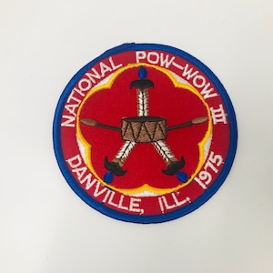 May include: A blue and red embroidered patch with the text "National Pow-Wow III Danville, Ill. 1975". The patch features a red background with a white outline and a yellow circle around the center. The center of the patch features a brown drum with three brown feathers on each side.