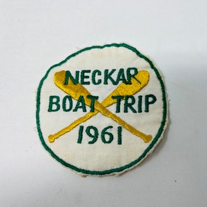 May include: A white embroidered patch with green stitching. The patch is circular and features two crossed oars with the text "Neckar Boat Trip 1961".