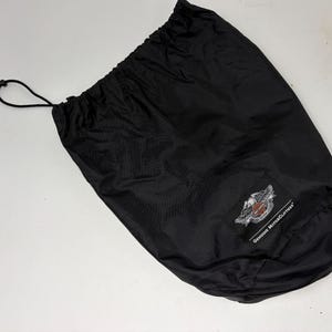 May include: Black drawstring bag with the Harley-Davidson logo on the lower right. The bag is made of a smooth, possibly nylon, material. The logo features a stylized eagle with the words "Genuine MotorClothes".