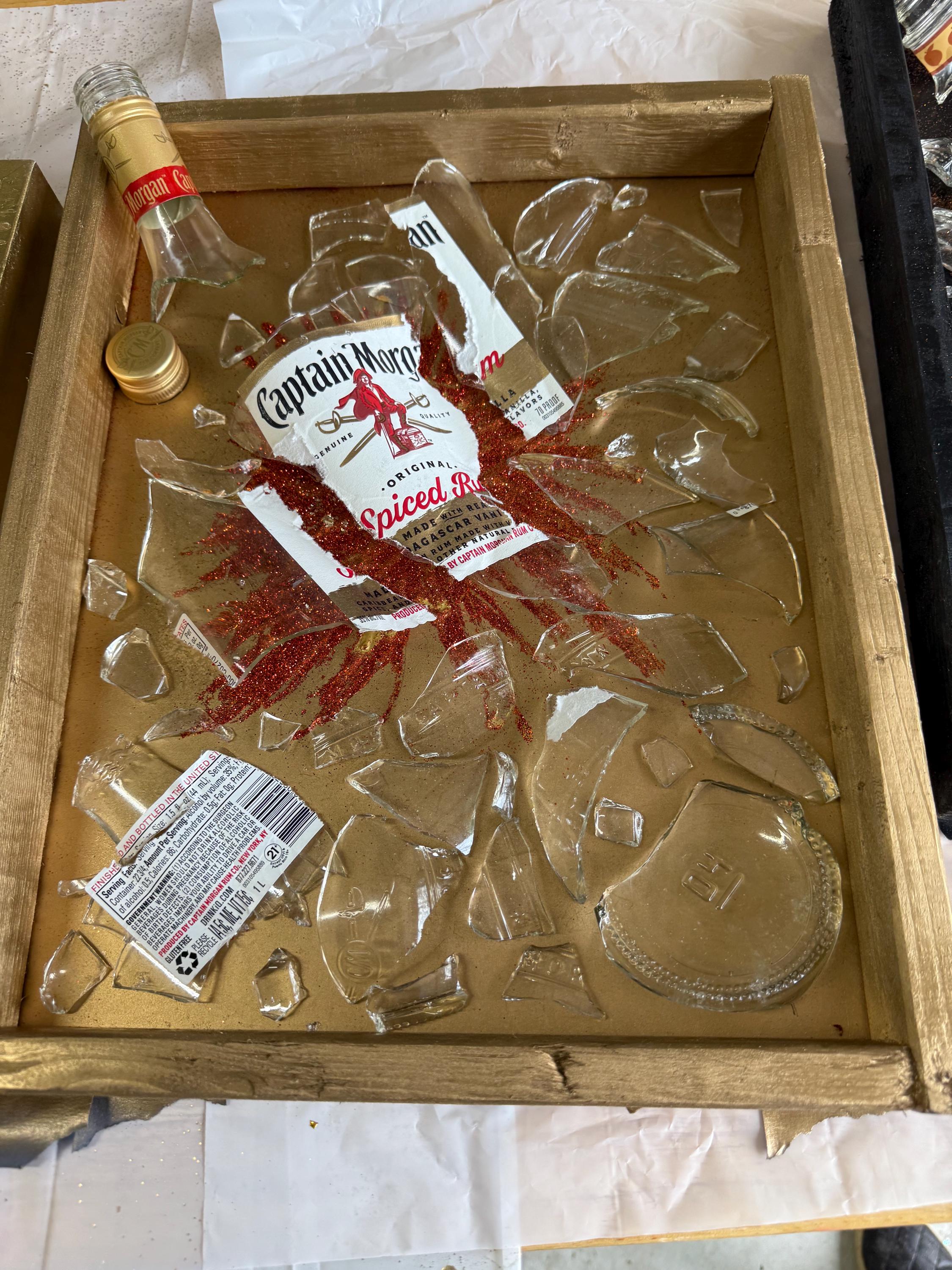 May include: A wooden frame with a gold background holds broken pieces of a glass bottle. The bottle label reads "Captain Morgan Original Spiced Rum". Red glitter is scattered around the broken glass.