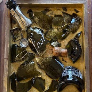 May include: A wooden box filled with broken pieces of a green glass champagne bottle, a cork, and gold glitter. The box is labeled "Lost".