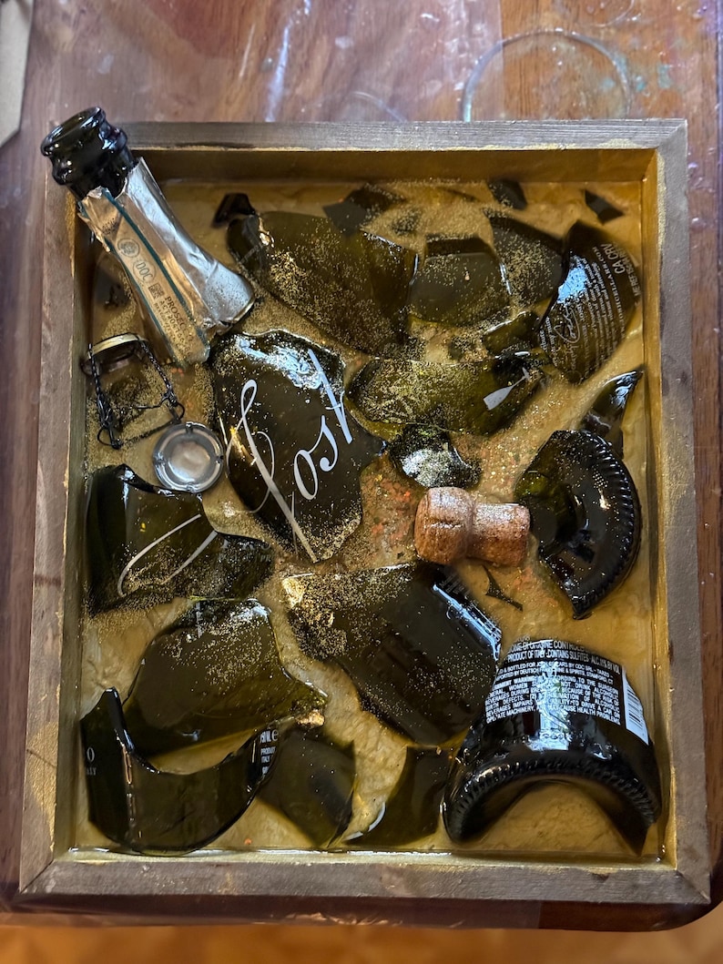 May include: A wooden box filled with broken pieces of a green glass champagne bottle, a cork, and gold glitter. The box is labeled "Lost".