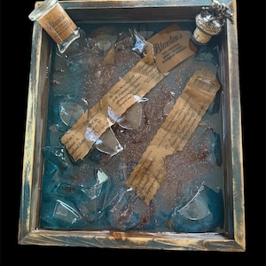 May include: A wooden tray with a blue resin interior containing broken glass pieces, a small bottle with a label that reads "Blanton's", and a cork stopper with a metal figure on top. The tray has a rustic, distressed look.