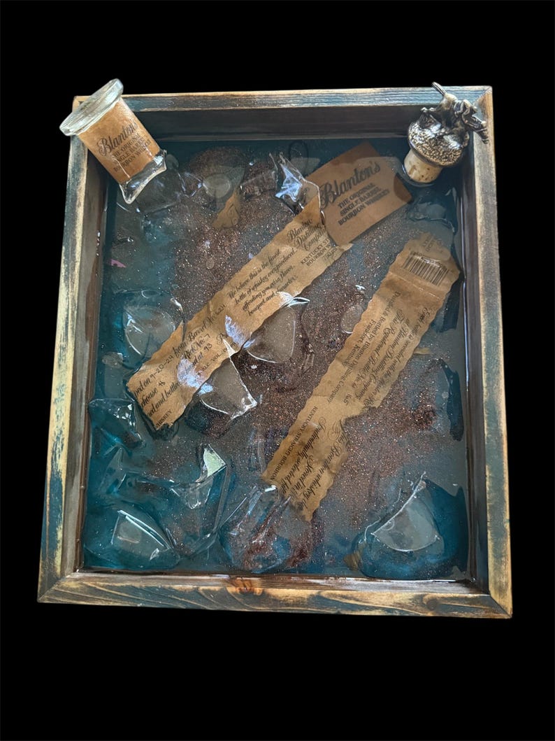 May include: A wooden tray with a blue resin interior containing broken glass pieces, a small bottle with a label that reads "Blanton's", and a cork stopper with a metal figure on top. The tray has a rustic, distressed look.