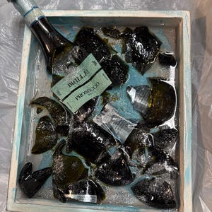 May include: A broken glass bottle of Prosecco, labeled "BRILLA", is submerged in a blue and white tray filled with glitter and water. The bottle is partially visible, with the label facing up.