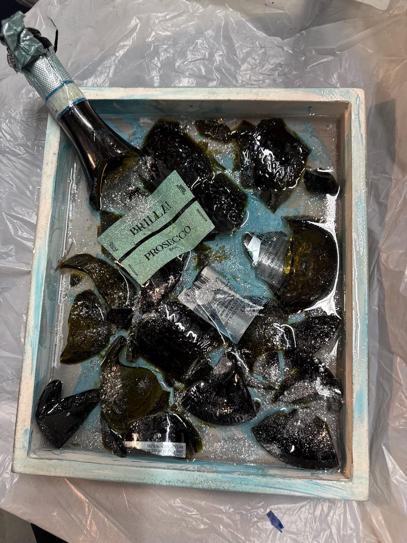 May include: A broken glass bottle of Prosecco, labeled "BRILLA", is submerged in a blue and white tray filled with glitter and water. The bottle is partially visible, with the label facing up.