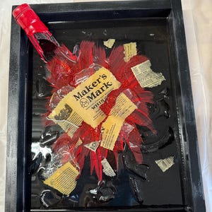 May include: A black framed resin art piece featuring a red Maker's Mark bourbon bottle broken into pieces with a red glitter background.