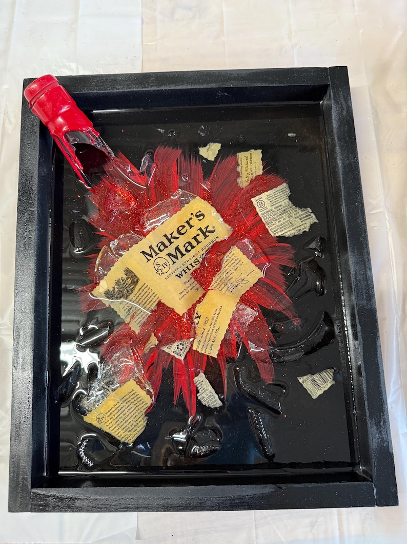 May include: A black framed resin art piece featuring a red Maker's Mark bourbon bottle broken into pieces with a red glitter background.