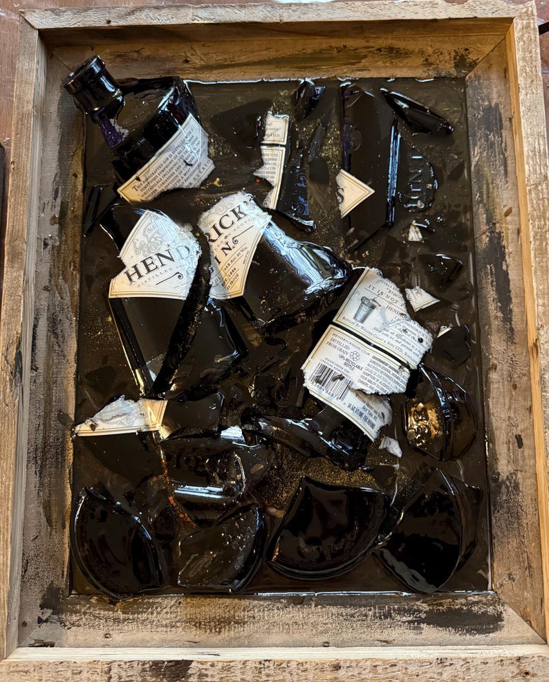 May include: A wooden frame holds a black resin art piece featuring broken glass bottles. The bottles are labeled "Hendrick's Gin" and "Tanqueray No. Ten Gin".