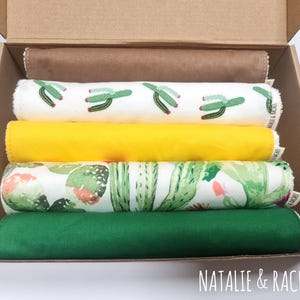 May include: A cardboard box contains four rolled fabric items. The top item is a brown fabric. Below are a white fabric with green cactus, a yellow fabric, a fabric with green cactus, and a green fabric. The text "NATALIE & RACHEL" is at the bottom.