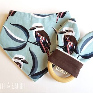 May include: A baby bib and teether set. The bib and teether are light blue with a pattern of kookaburras and leaves in brown, white, and blue. The teether has a wooden ring and a brown fabric accent. The brand name "NATALIE & RACHEL" is printed on the teether.