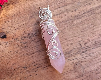 May include: A pink rose quartz crystal pendant wrapped in silver wire. The pendant is on a wooden surface with pink flowers.