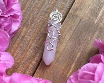 May include: A pink crystal pendant wrapped in silver wire. The pendant is surrounded by pink carnations on a wooden background.