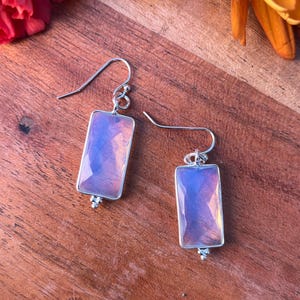 Może przedstawiać: Para prostokątnych kolczyków ze srebrną oprawą i jasno różowym, opalizującym kamieniem. Kolczyki są prezentowane na drewnianej powierzchni z pomarańczowymi i czerwonymi kwiatami.