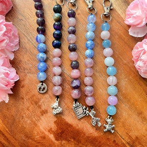 Puede incluir: Cinco llaveros de cuentas con cuentas de diferentes colores y dijes. Los dijes incluyen un osito de peluche, un cochecito, una huella y un certificado con el texto "Birth Certificate Date: Time: Weight: Length:".