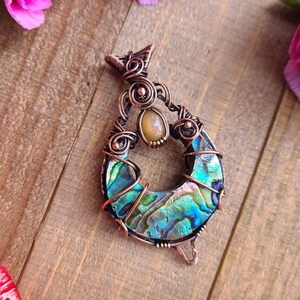 May include: A handcrafted copper pendant with an abalone shell inlay, featuring iridescent blues, greens, and purples. The pendant is accented with a small, round, yellow gemstone. Pink carnations and a wooden surface provide the background.
