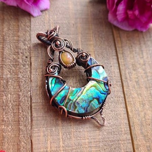 May include: A handcrafted copper pendant with an abalone shell inlay, featuring iridescent blues and greens. The pendant is wire-wrapped and includes a small, oval-shaped stone. Pink carnations are scattered around the pendant on a wooden surface.