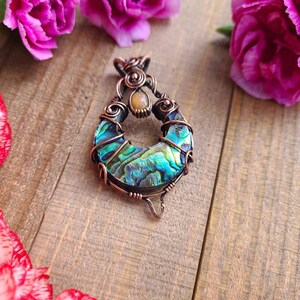May include: A handcrafted pendant featuring a vibrant abalone shell set in copper wire. The pendant has a teardrop shape with iridescent blues, greens, and purples. A small, round, light-colored stone is set at the top. Pink flowers are in the background.