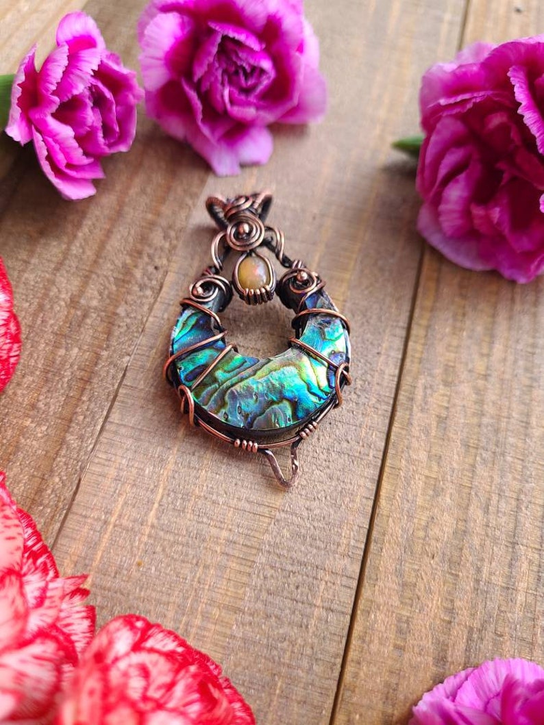 May include: A handcrafted pendant featuring a vibrant abalone shell set in copper wire. The pendant has a teardrop shape with iridescent blues, greens, and purples. A small, round, light-colored stone is set at the top. Pink flowers are in the background.