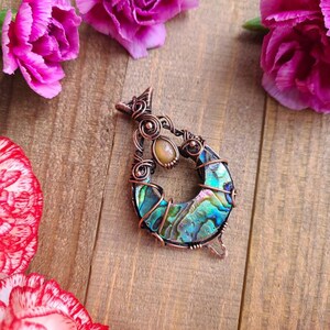 May include: A handcrafted pendant with a copper wire frame, featuring a vibrant abalone shell crescent and a small, oval-shaped stone. The pendant is set against a wooden background, surrounded by pink carnation flowers.