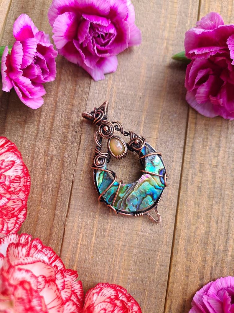 May include: A handcrafted pendant with a copper wire frame, featuring a vibrant abalone shell crescent and a small, oval-shaped stone. The pendant is set against a wooden background, surrounded by pink carnation flowers.