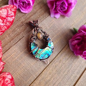 May include: A handcrafted copper pendant with an abalone shell inlay and a small, round, light-colored stone. The pendant is surrounded by pink carnations on a wooden surface. The abalone shell displays iridescent blues and greens.