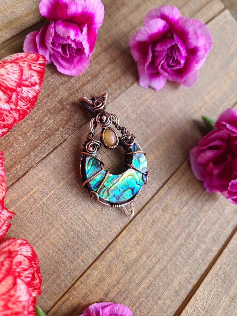 May include: A handcrafted copper pendant with an abalone shell inlay and a small, round, light-colored stone. The pendant is surrounded by pink carnations on a wooden surface. The abalone shell displays iridescent blues and greens.