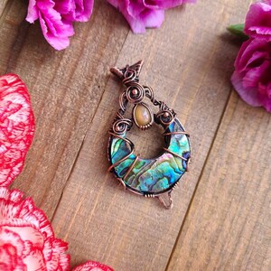 May include: A handcrafted copper pendant with an abalone shell inlay, featuring iridescent blues, greens, and purples. The pendant is surrounded by intricate wirework and a small, oval-shaped stone. Pink carnations are arranged around the pendant on a wooden surface.
