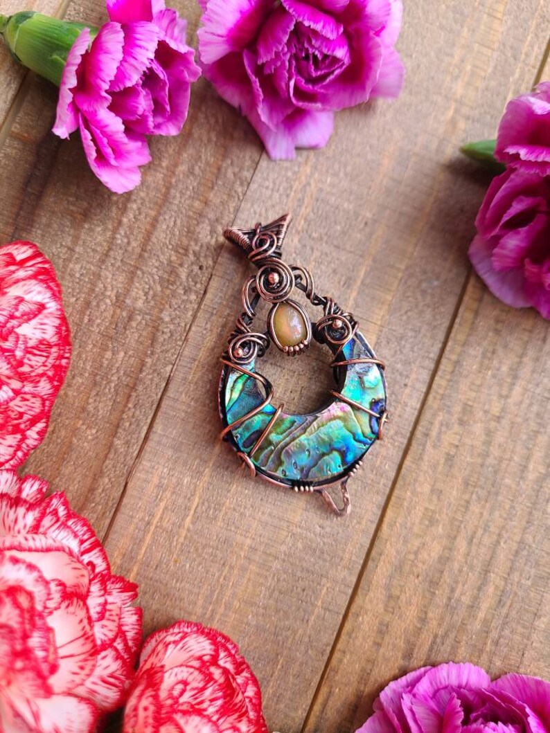 May include: A handcrafted copper pendant with an abalone shell inlay, featuring iridescent blues, greens, and purples. The pendant is surrounded by intricate wirework and a small, oval-shaped stone. Pink carnations are arranged around the pendant on a wooden surface.