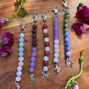 May include: Six colorful beaded keychains with different charms. The charms include a baby bottle, a stroller, a rocking horse, a teddy bear, a book with the text "Happy Birthday!" and a green and blue charm.