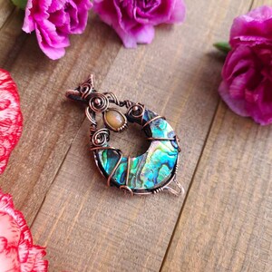 May include: A handcrafted pendant featuring a crescent-shaped abalone shell, wrapped in copper wire. The pendant includes a small, oval-shaped stone and is surrounded by pink carnation flowers on a wooden surface.