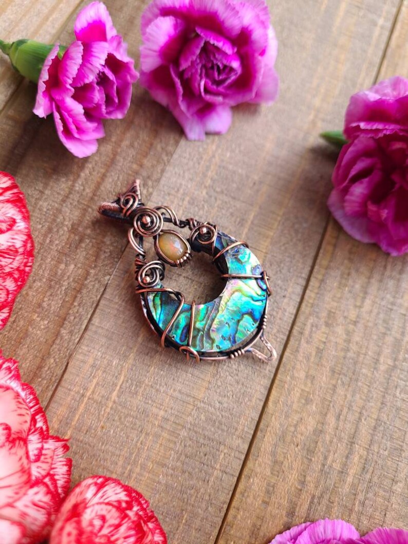 May include: A handcrafted pendant featuring a crescent-shaped abalone shell, wrapped in copper wire. The pendant includes a small, oval-shaped stone and is surrounded by pink carnation flowers on a wooden surface.