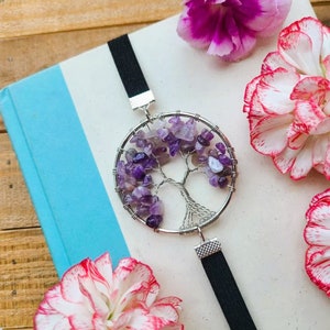 May include: A purple amethyst wire wrapped tree of life bookmark with a black elastic strap. The bookmark is on a blue and white book with pink flowers.