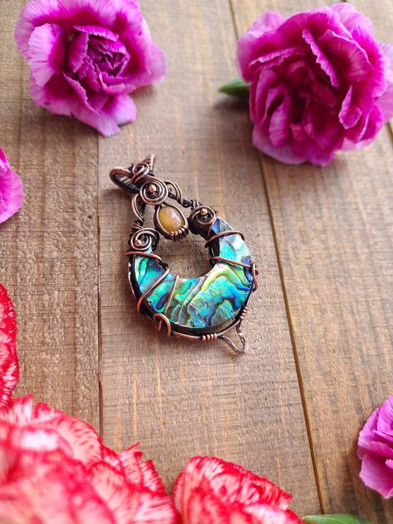 May include: A handcrafted copper pendant with an abalone shell inlay, featuring iridescent blues and greens. The pendant is wire-wrapped and includes a small, oval-shaped stone. Pink carnations are scattered around the pendant on a wooden surface.