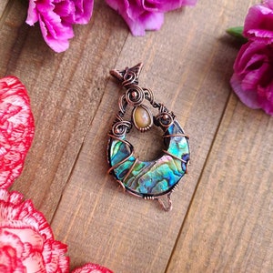 May include: A handcrafted copper pendant with an abalone shell inlay, showcasing iridescent blues, greens, and purples. The pendant is adorned with a small, oval-shaped stone and intricate wire wrapping. Pink carnations surround the pendant on a wooden surface.